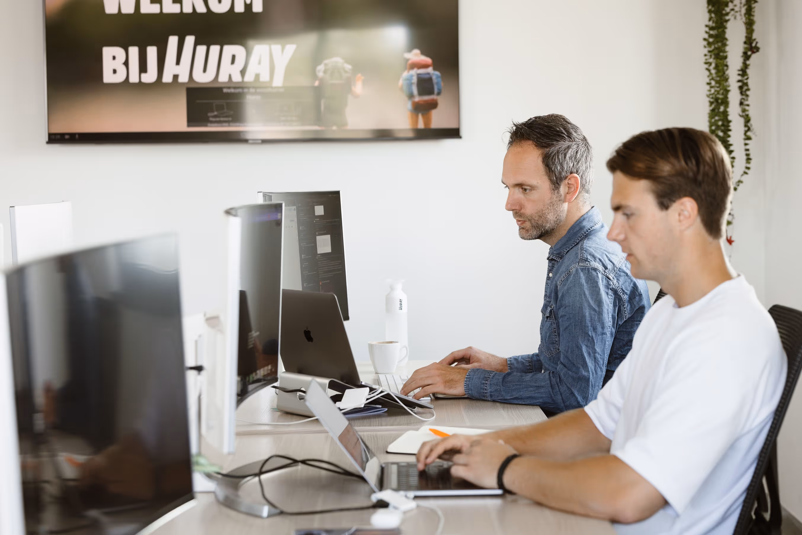 Twee mannen werken geconcentreerd op laptops aan een bureau in een modern kantoor.