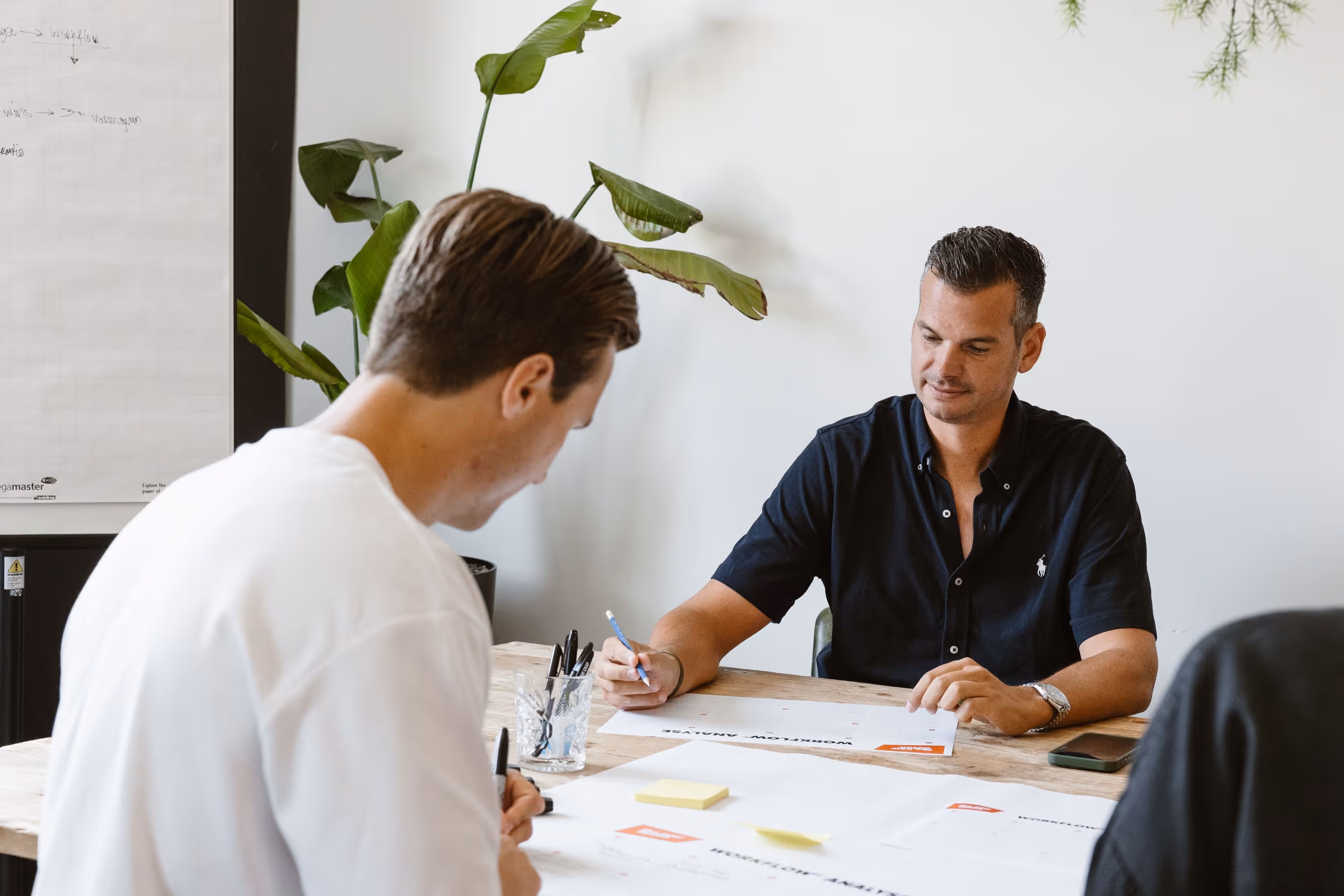 Twee mannen zitten aan tafel en bekijken documenten met notities tijdens een vergadering.
