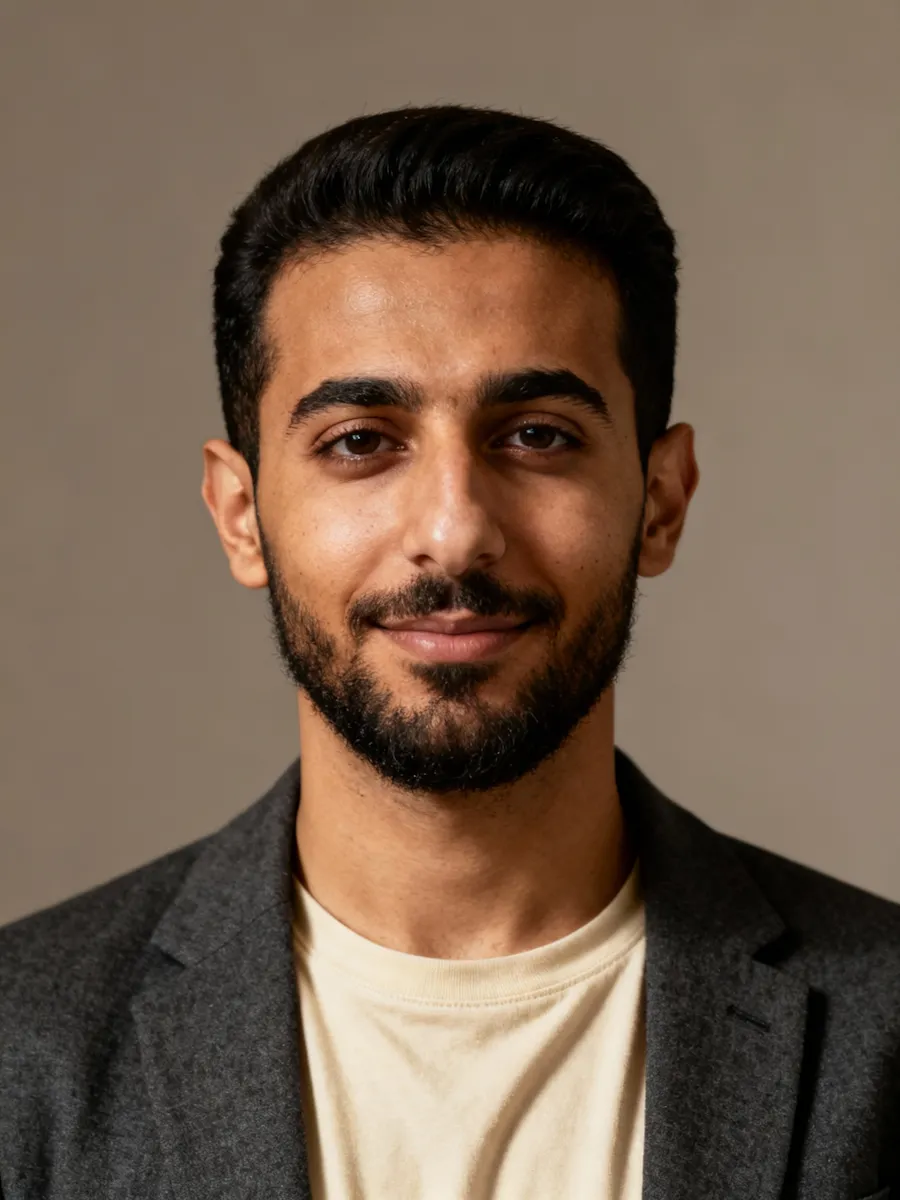 Smiling man with short dark hair and beard wearing a light shirt and dark blazer against a neutral background.