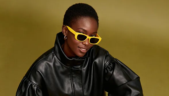 Smiling woman wearing yellow sunglasses and a black leather jacket against an olive background.
