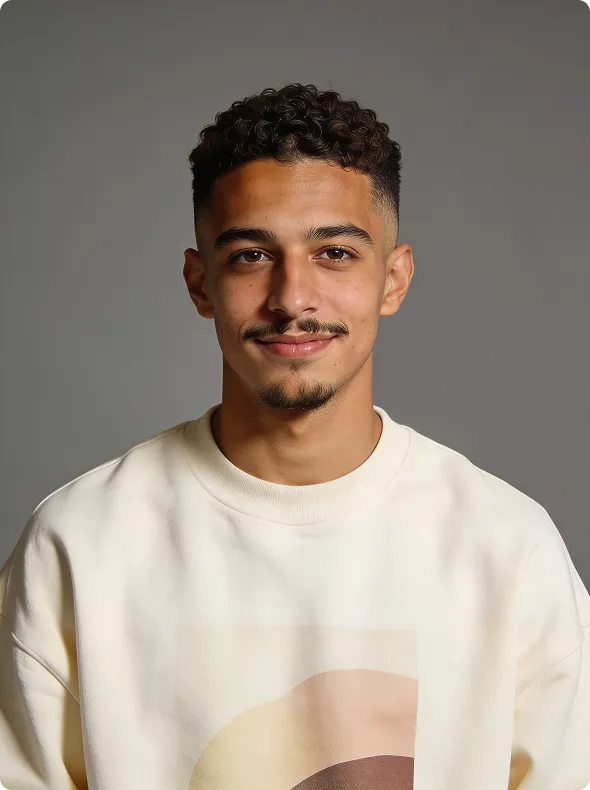 Young man with curly hair and mustache smiles gently, wearing a cream sweatshirt against a gray background.