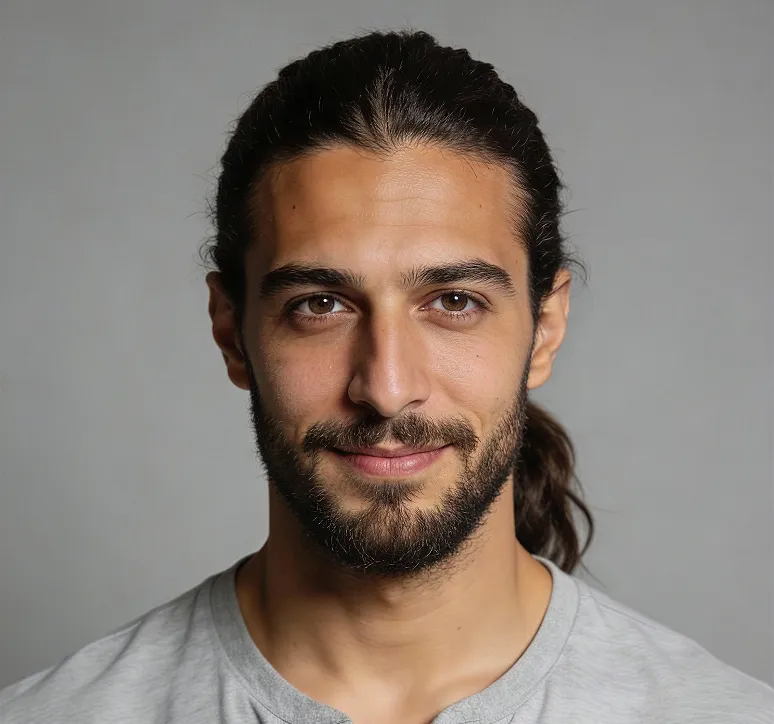 Smiling man with dark hair tied back and beard wearing a light gray shirt against a plain gray background.