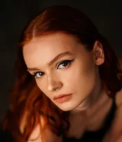Close-up portrait of a young red-haired woman with winged eyeliner and a neutral expression.