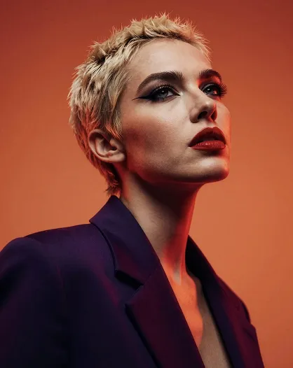 Portrait of a woman with short blonde hair, bold makeup, and a dark blazer against an orange background.