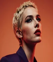 Portrait of a person with short platinum blond hair and bold makeup against an orange background.