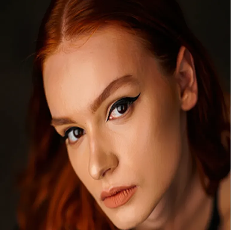 Close-up of a woman with red hair, winged eyeliner, and muted orange lipstick against a dark background.