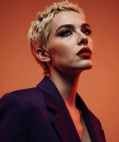 Portrait of a person with short blonde hair, dark makeup, and wearing a dark jacket against orange background.