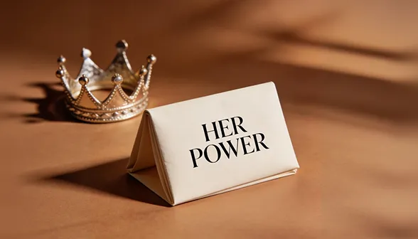 A small crown sits next to a folded cream-colored card that reads 'HER POWER' on a brown surface.
