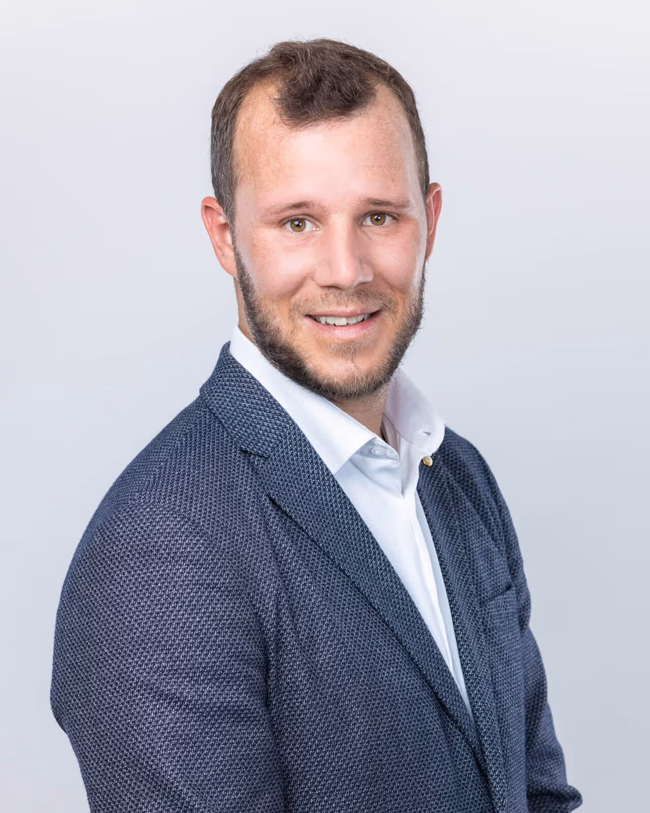 Homme souriant en costume bleu foncé et chemise blanche sur fond gris clair.