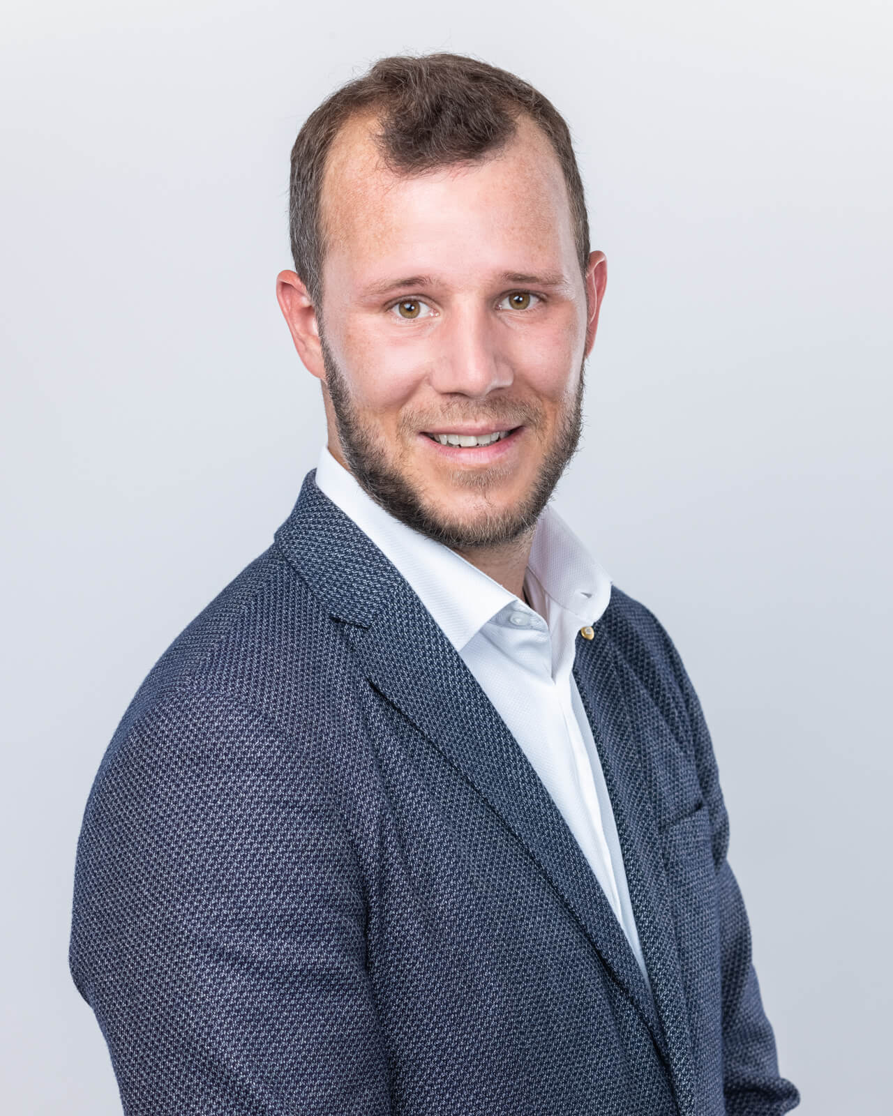 Homme souriant en costume bleu foncé et chemise blanche sur fond gris clair.