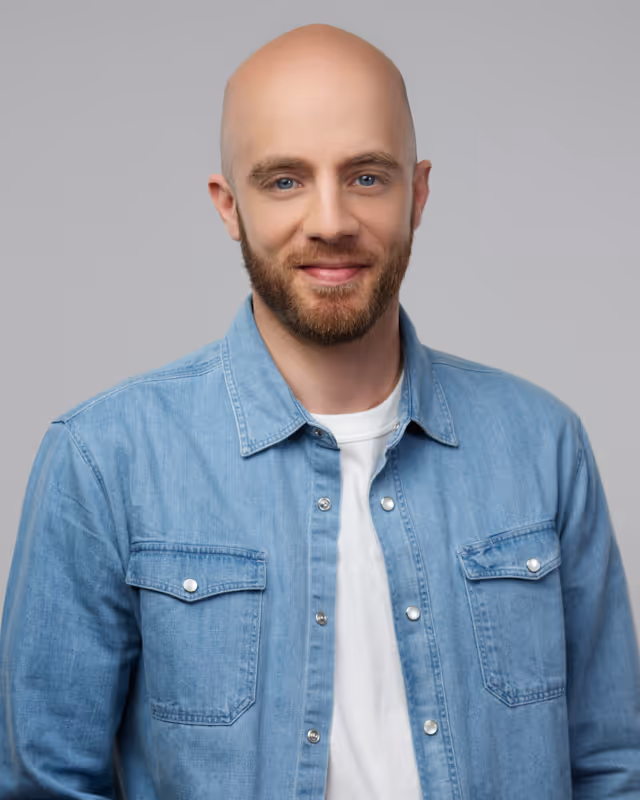 Homme souriant avec une barbe, portant une chemise en jean bleu sur un t-shirt blanc.