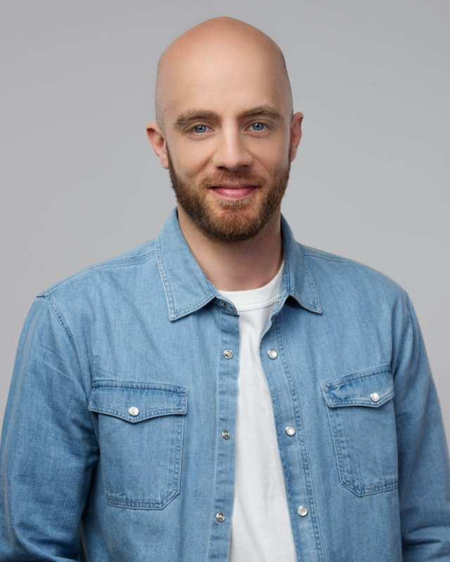 Homme souriant avec une barbe, portant une chemise en jean bleu sur un t-shirt blanc.