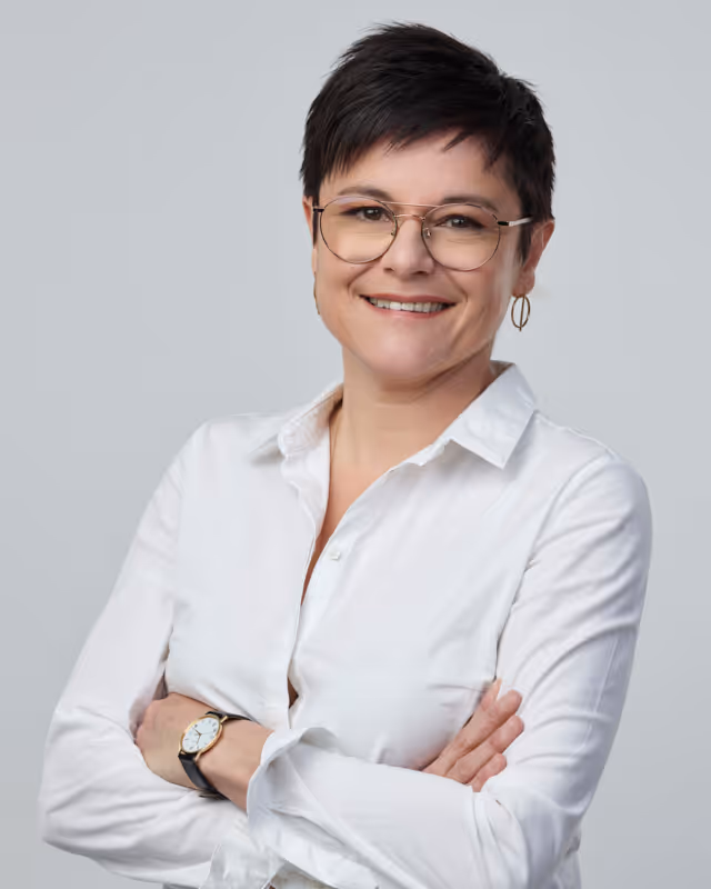 Femme souriante portant des lunettes et une chemise blanche, bras croisés devant un fond gris clair.