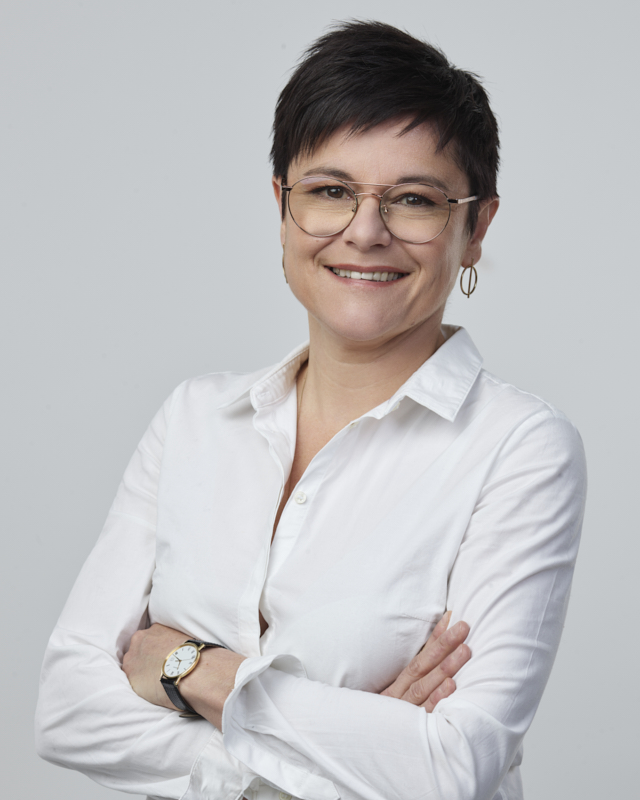 Femme souriante portant des lunettes et une chemise blanche, bras croisés devant un fond gris clair.