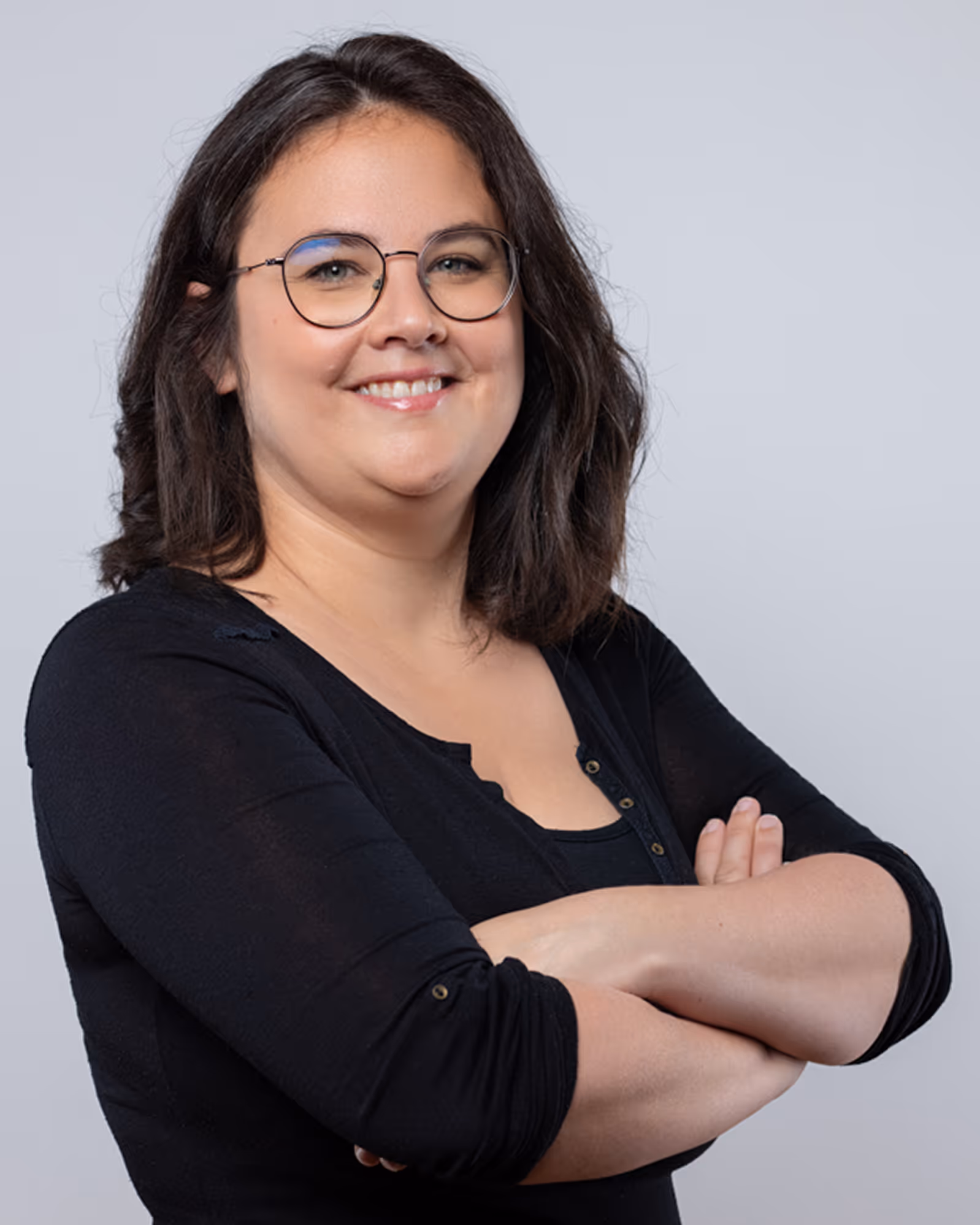 Une femme aux cheveux bruns ondulés, portant des lunettes rondes et un haut noir, souriante bras croisés.