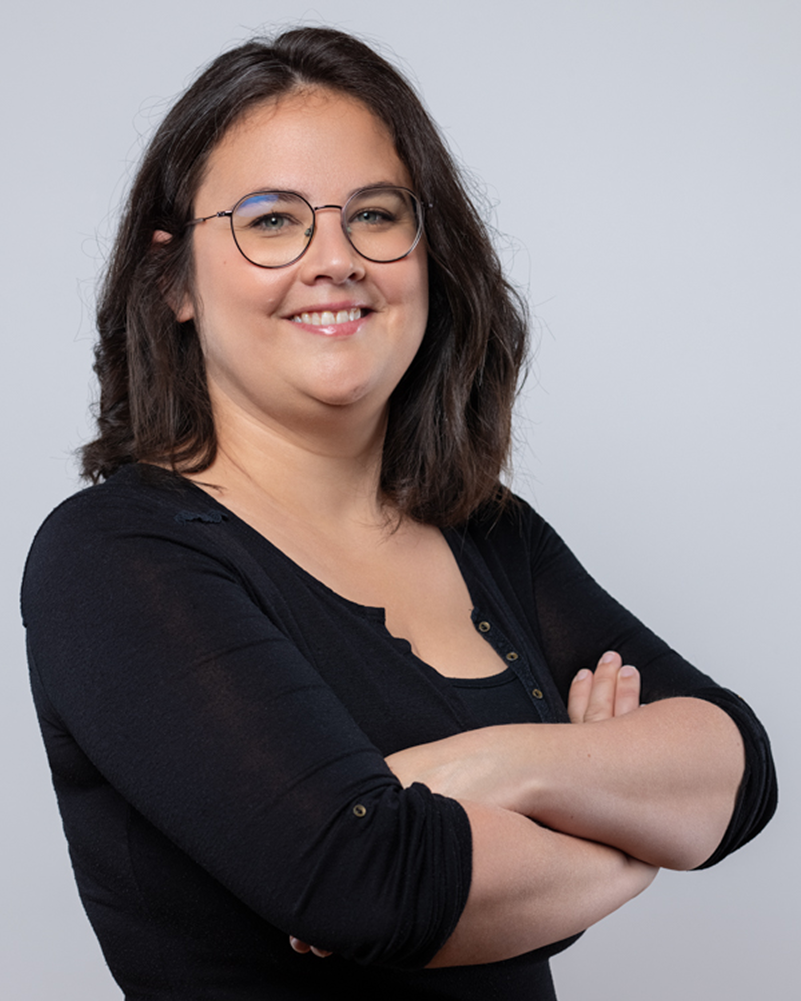Une femme aux cheveux bruns ondulés, portant des lunettes rondes et un haut noir, souriante bras croisés.