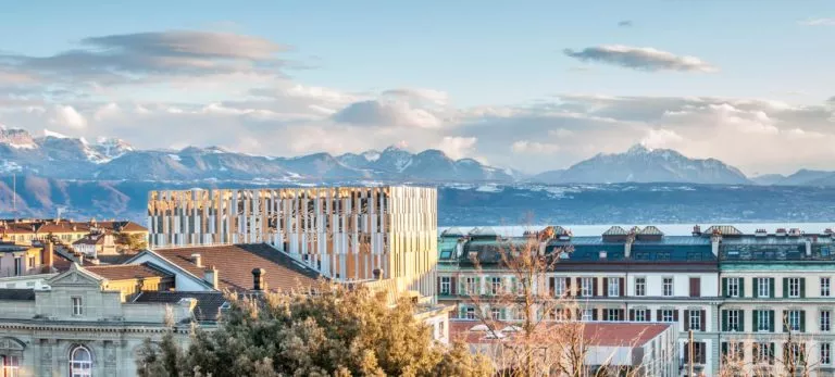 Vue urbaine avec immeubles modernes et montagnes enneigées en arrière-plan sous un ciel partiellement nuageux.