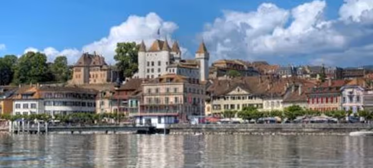 Vue de la ville au bord de l'eau avec des bâtiments anciens et un château sous un ciel bleu avec des nuages.