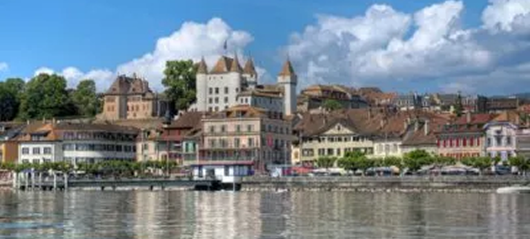 Vue de la ville au bord de l'eau avec des bâtiments anciens et un château sous un ciel bleu avec des nuages.