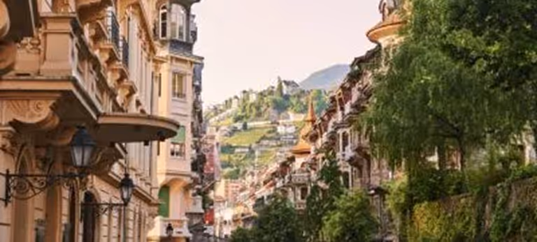 Rue pittoresque bordée de bâtiments anciens avec une colline verte et des maisons en arrière-plan.