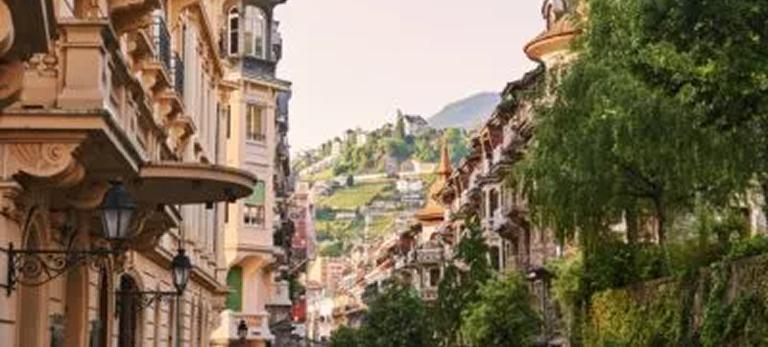 Rue pittoresque bordée de bâtiments anciens avec une colline verte et des maisons en arrière-plan.
