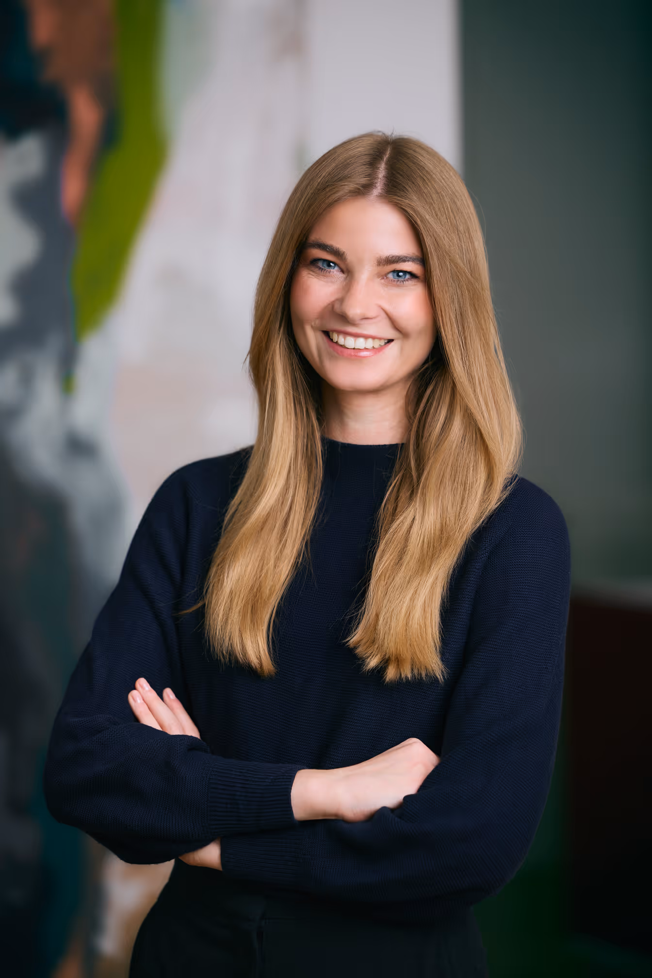 Fröhliche junge Frau mit langen blonden Haaren und blauem Pullover, Arme verschränkt, vor unscharfem Hintergrund.