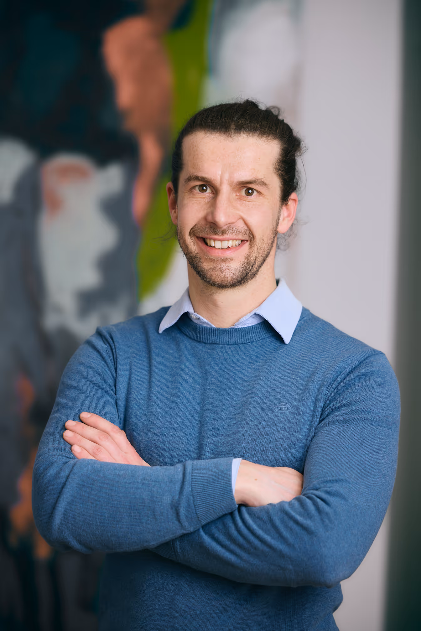 Lächelnder Mann mit zusammengelegten Armen trägt blauen Pullover und Hemd, unscharfer Hintergrund.