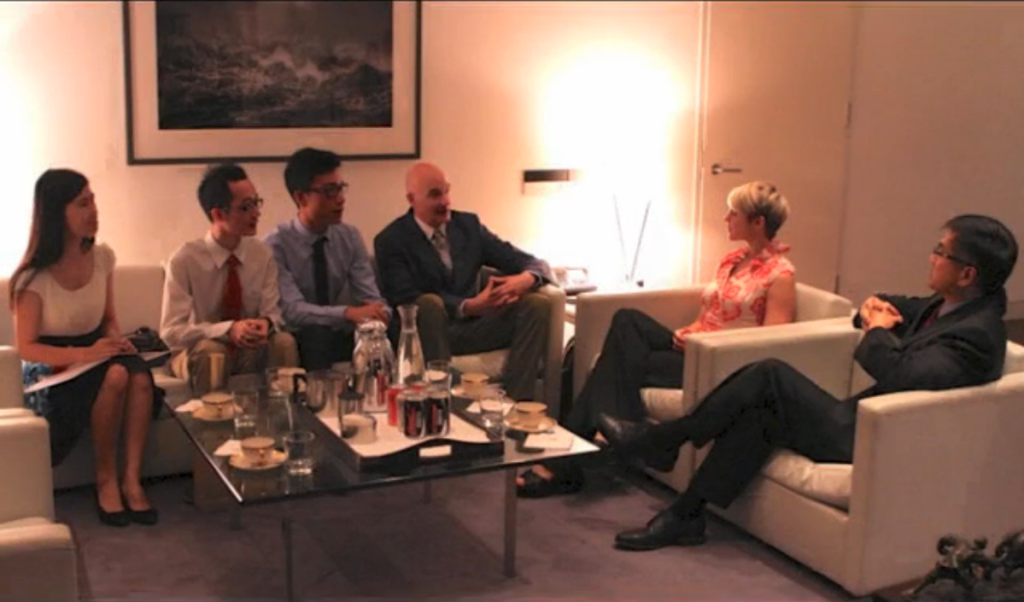 A group in business attire sit in a meeting with refreshments on a table.