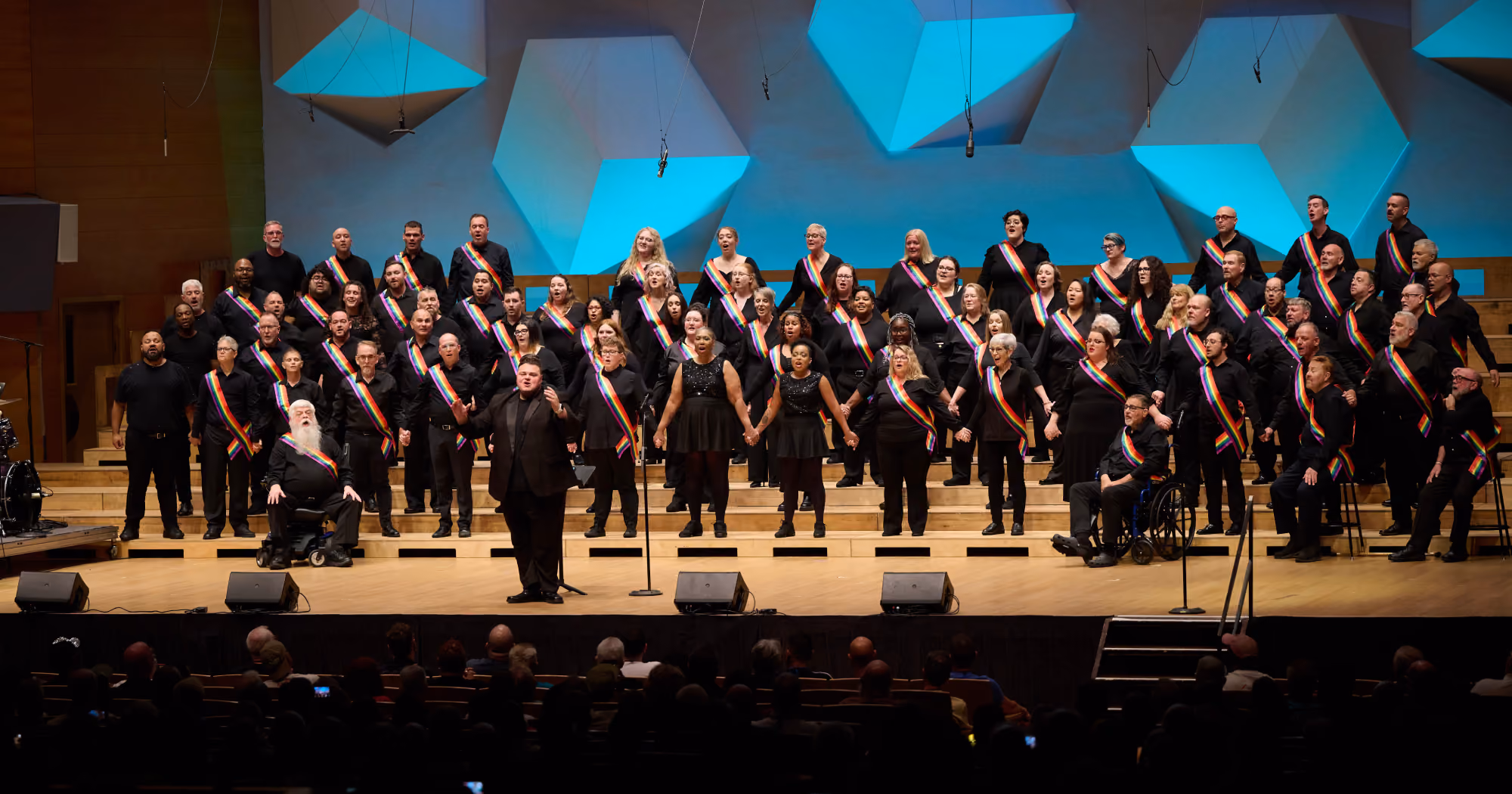 The Orlando Gay Chorus sing on stage hand in hand with rainbow sashes over their chests, facing the audience with their conductor
