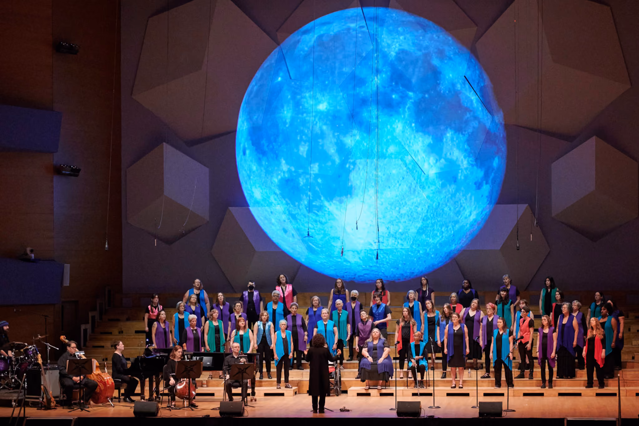 The ANNA Crusis Feminist Chorus performs on stage at GALA Festival 2024, with a large projection of the moon looming behind them