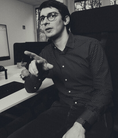 Man wearing glasses and a patterned shirt sitting at a desk, pointing forward with an engaged expression.