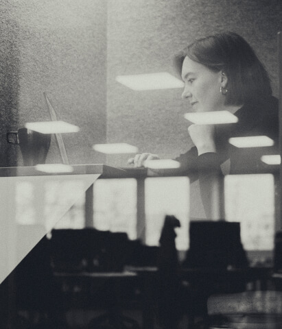 Reflection of a woman looking thoughtfully at a laptop in a dimly lit office with ceiling lights and windows visible.