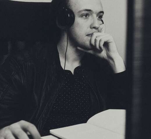 Young man wearing headphones, looking thoughtfully at a computer screen with an open book in front of him.