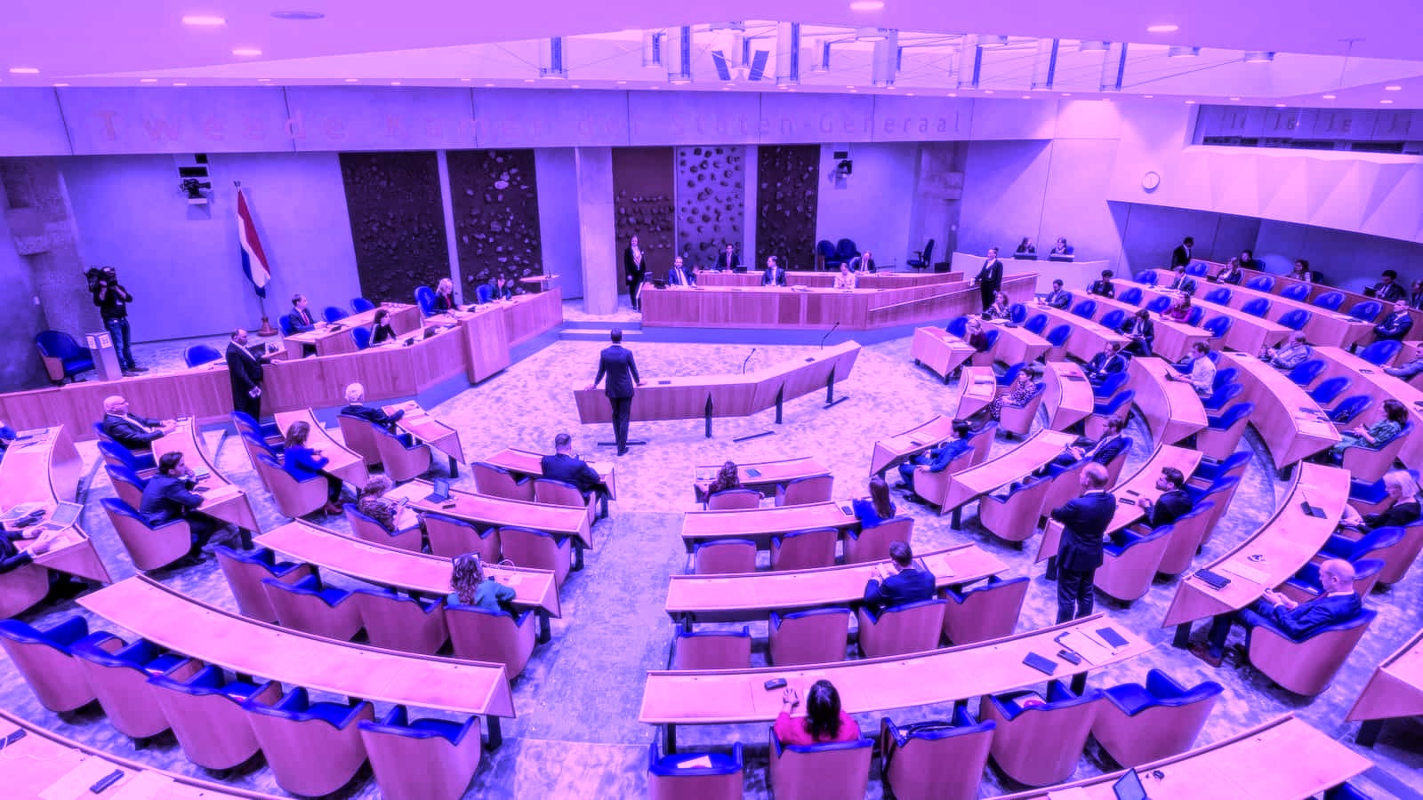 Plenary chamber with semicircular rows of seats and a Dutch flag, seen from the gallery