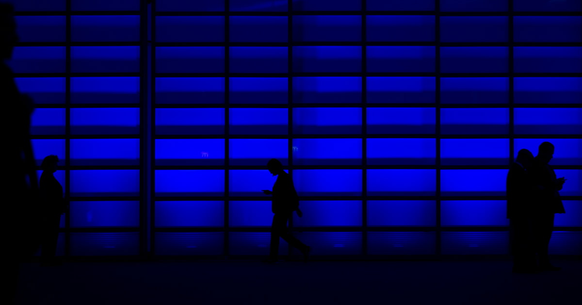 Silhouetten van mensen voor een blauw verlichte glazen gevel, met een man die lopend op zijn telefoon kijkt