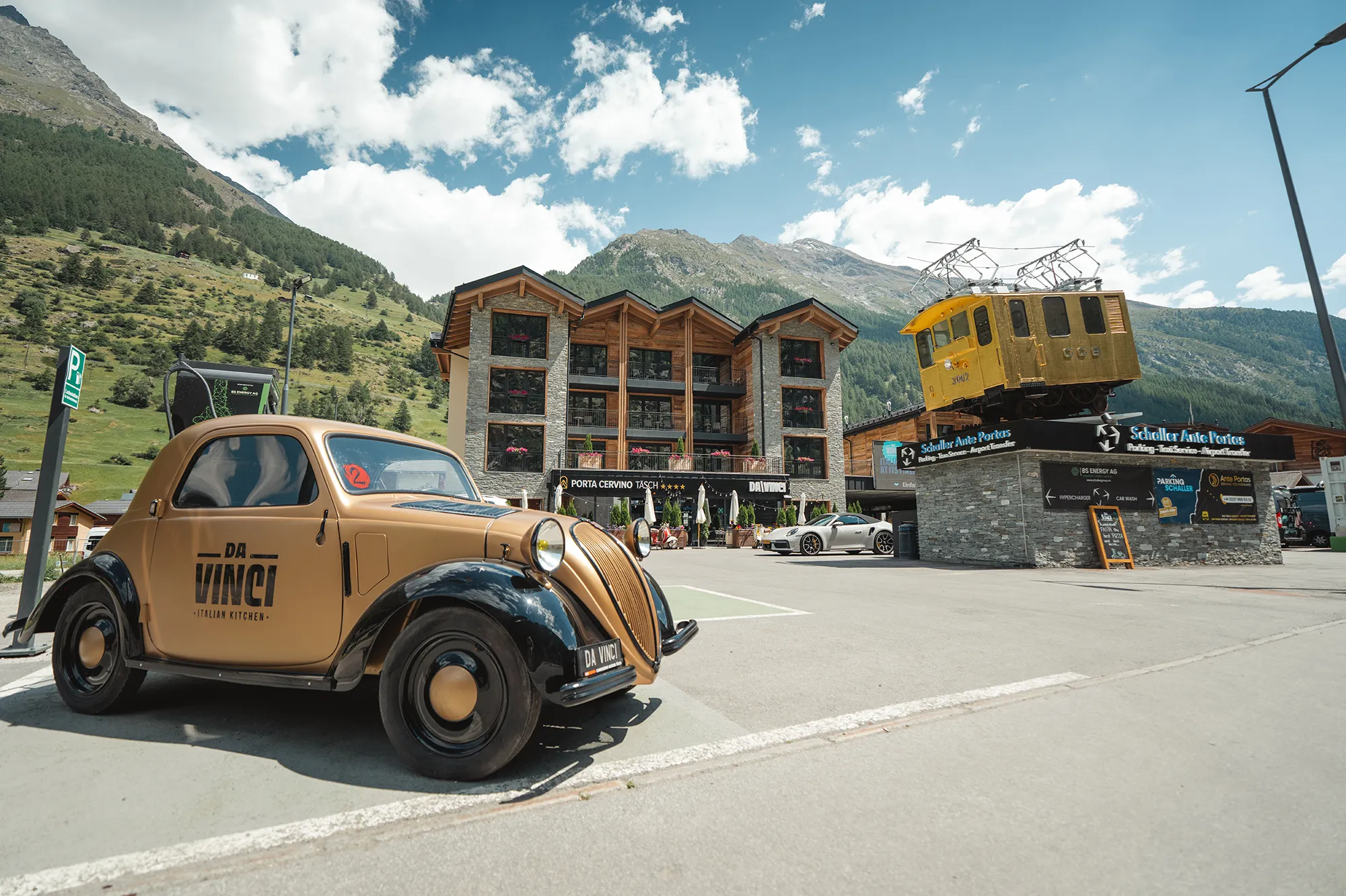 Hotel Porta Cervino in Täsch, featuring the Da Vinci restaurant, a golden vintage car, and the golden locomotive at the Schaller parking lot