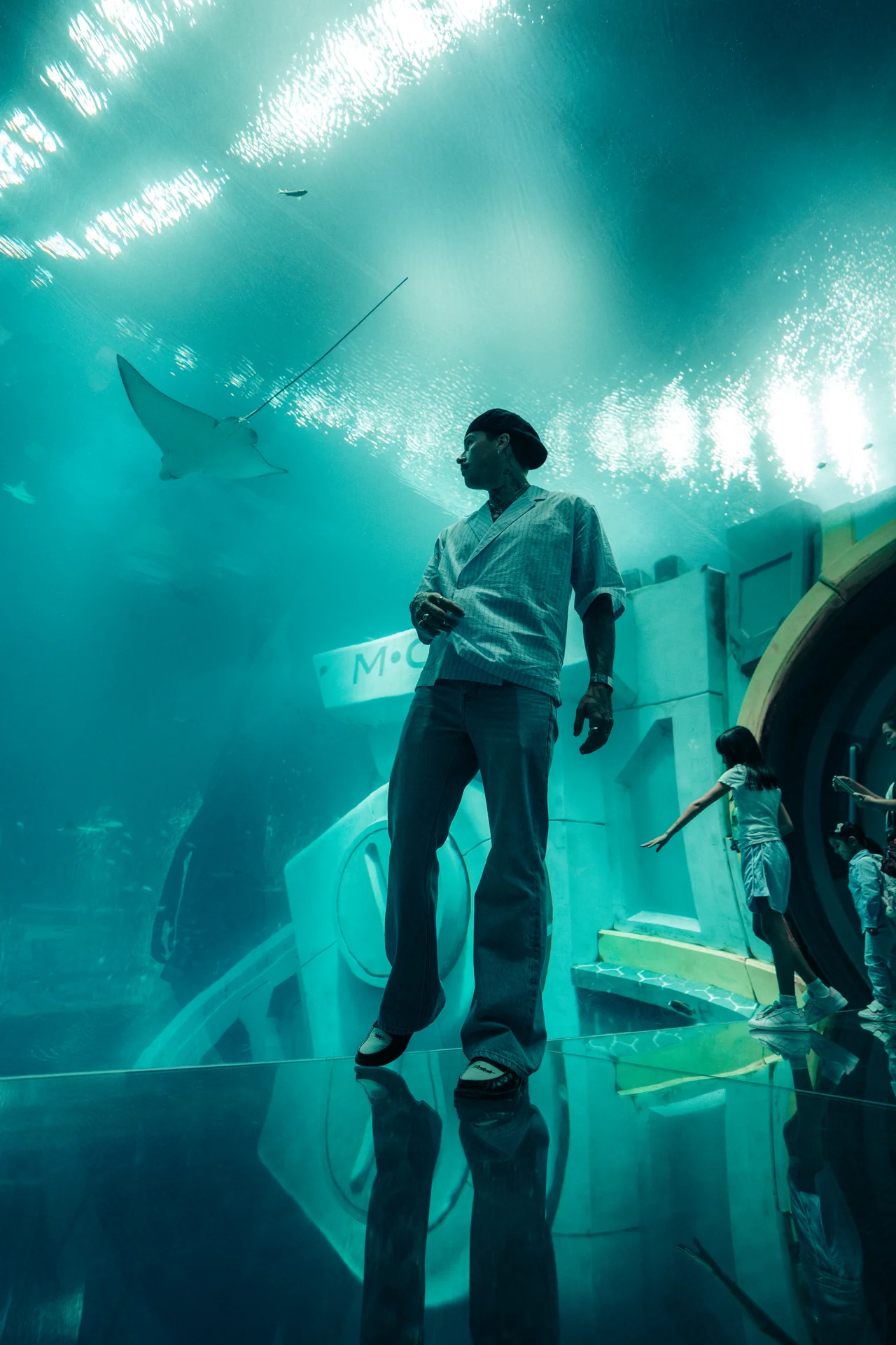 Man in casual clothes standing inside an aquarium tunnel observing a stingray swimming overhead.