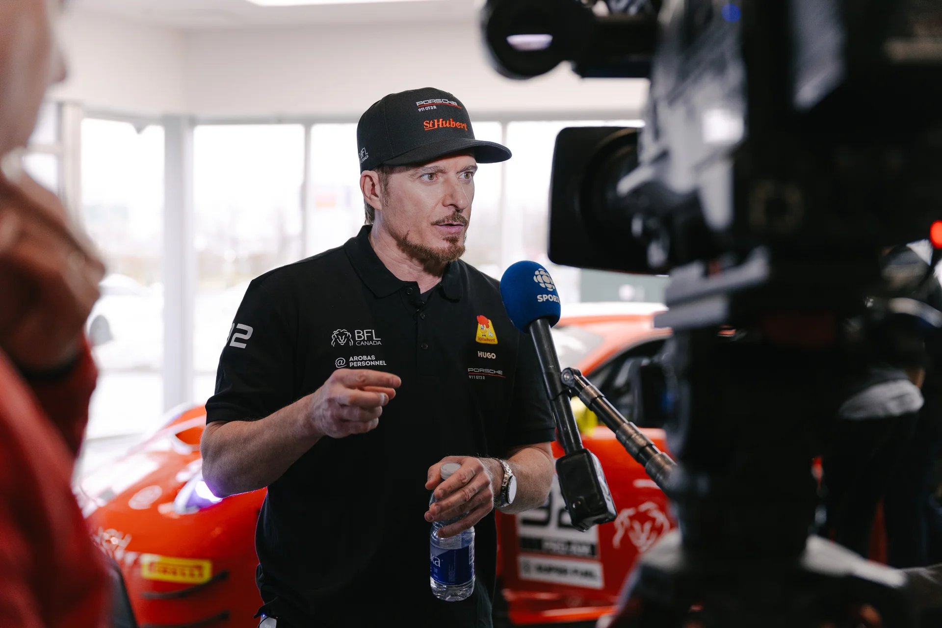 Man in black Porsche racing shirt and cap holding water bottle being interviewed by CBC Sports in front of a red race car.