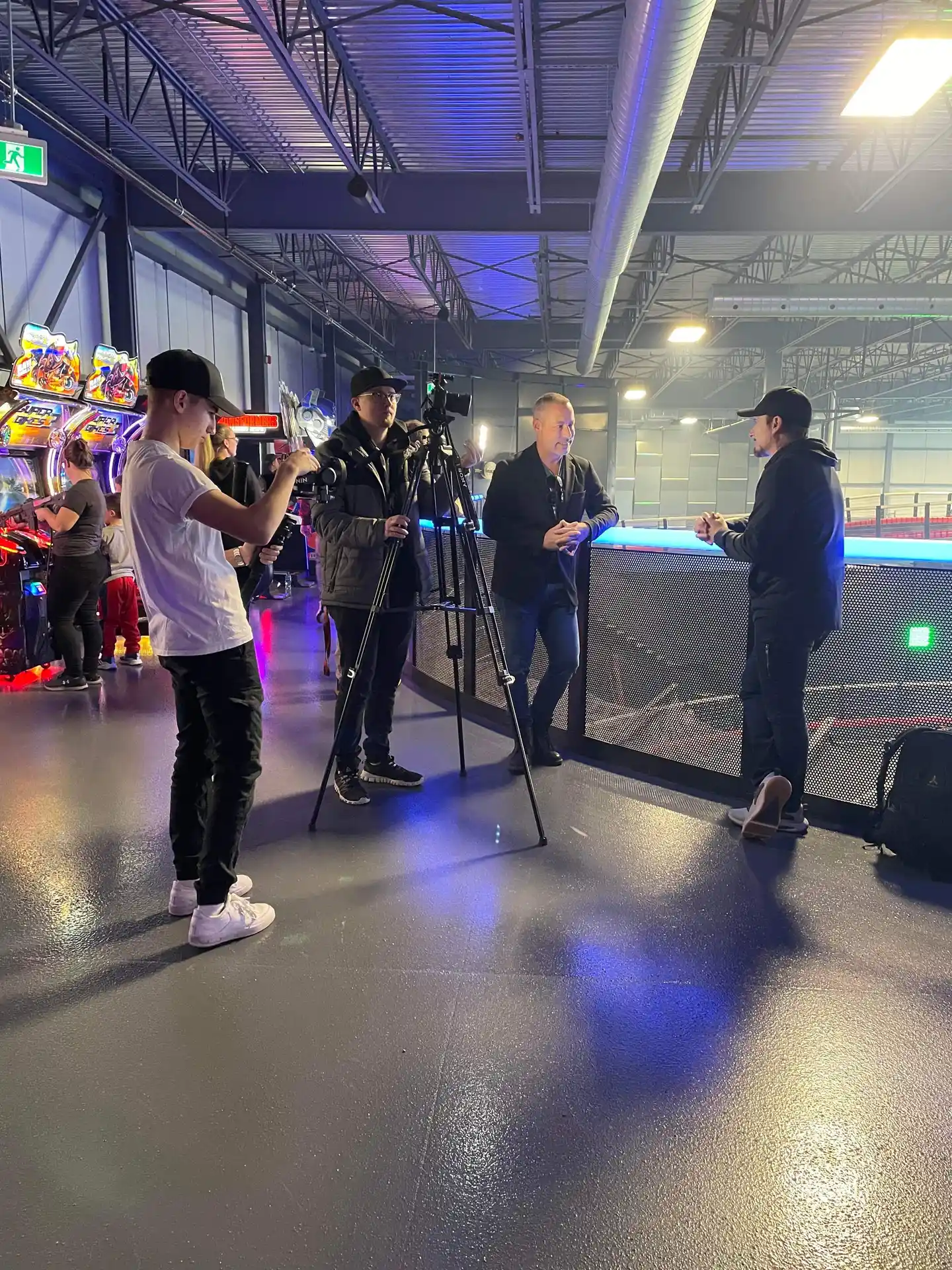 Two men having an interview near a railing inside an arcade with cameras and a cameraman recording.