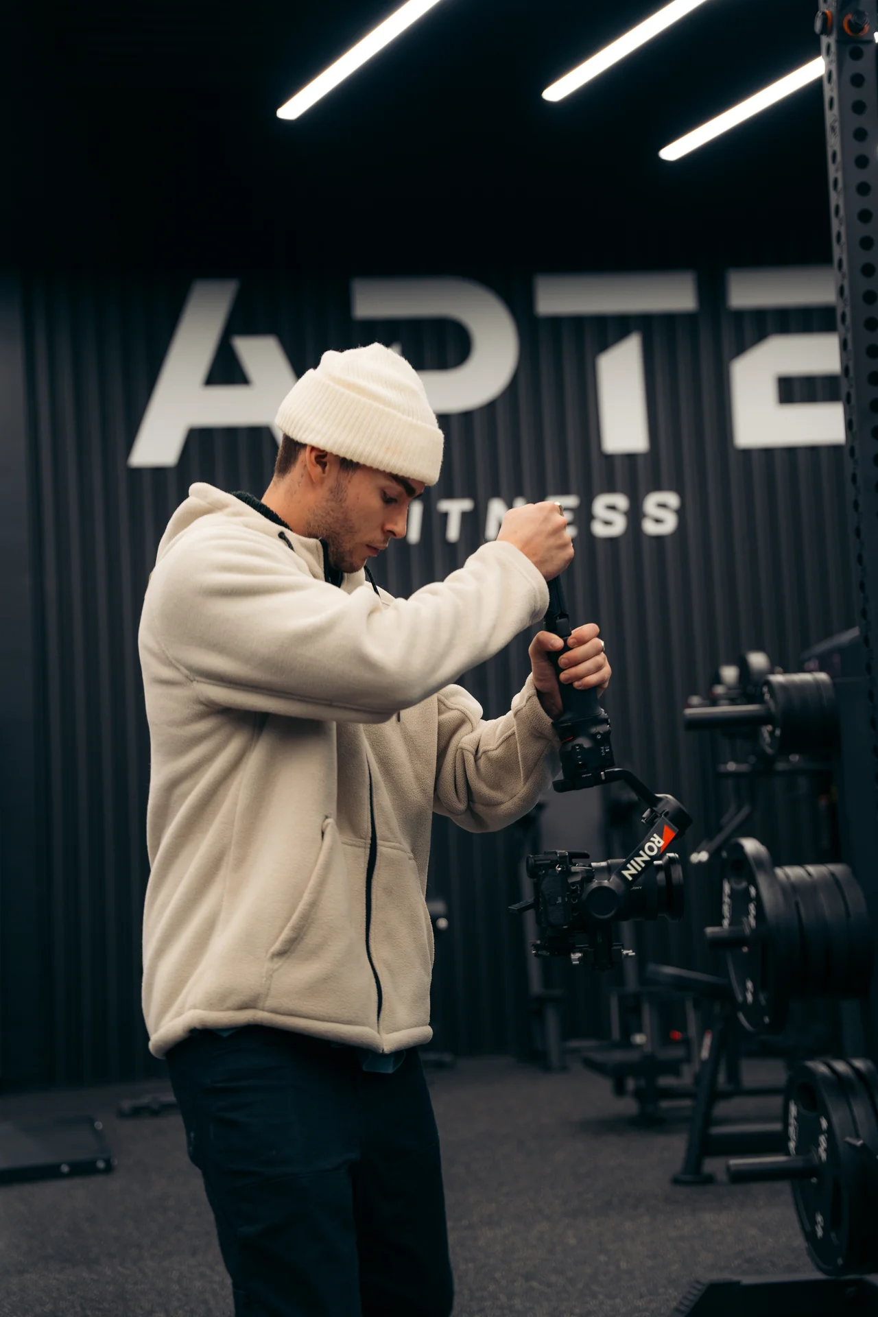 Man in beige jacket and beanie adjusting a Ronin camera stabilizer in a gym with weights.