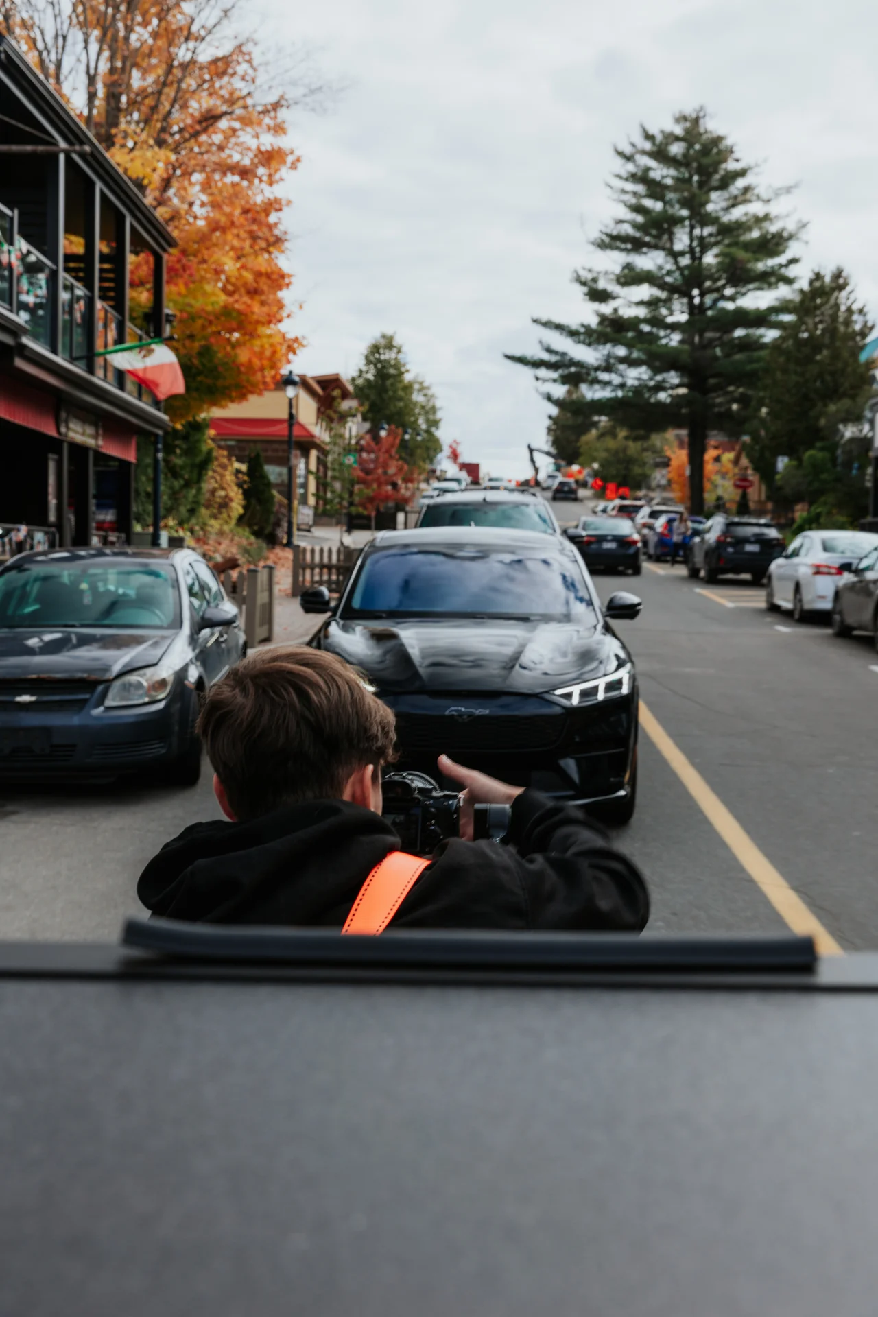 Person with camera taking a photo of a black Mustang car on a street with autumn trees.