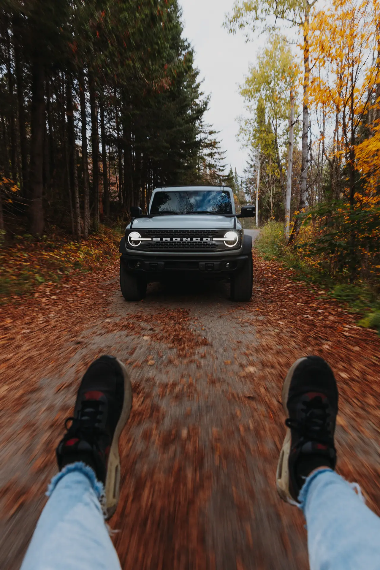 Front view of a Ford Bronco driving on a leaf-covered forest path with person's legs and shoes visible.