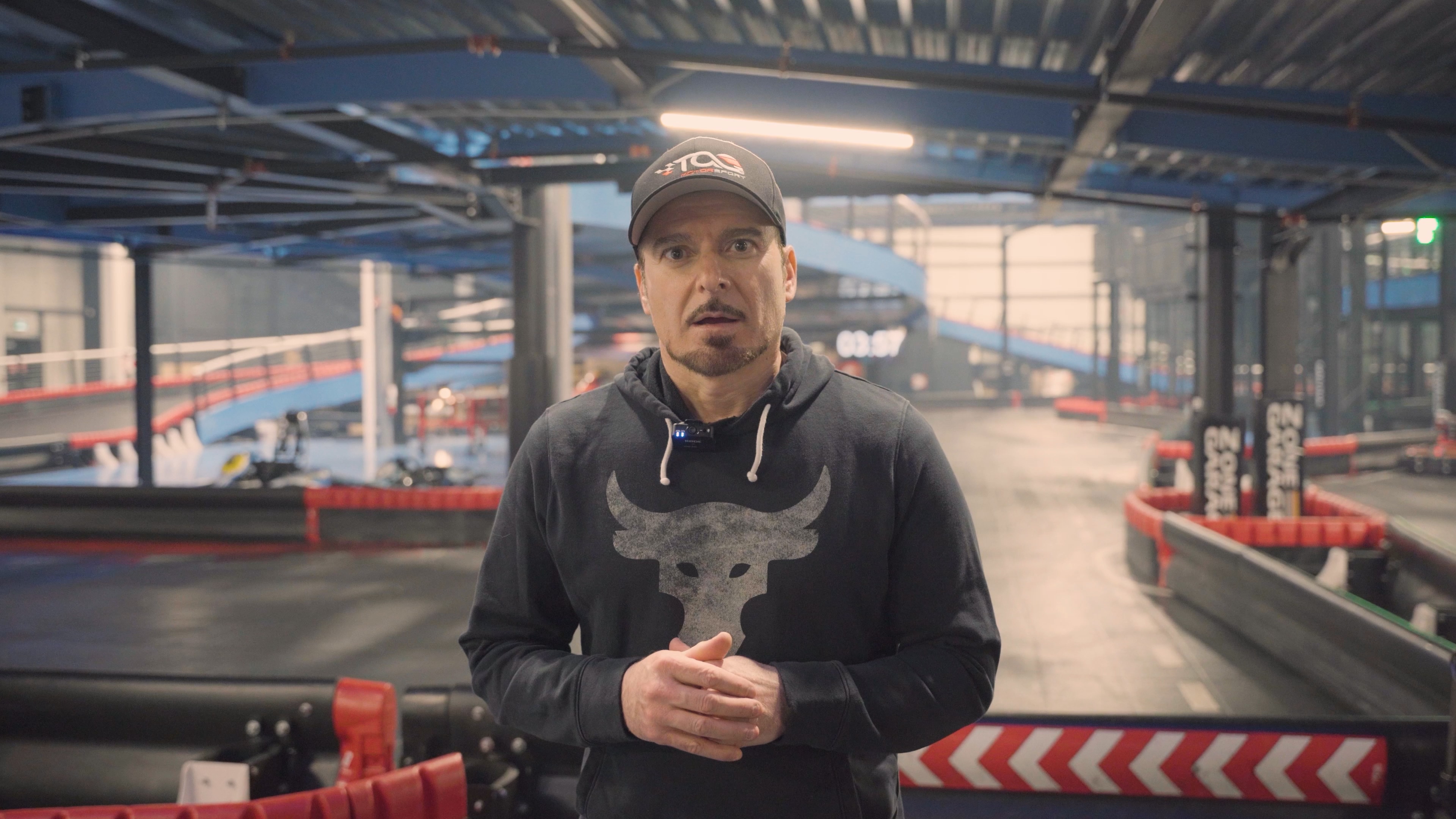 Man wearing a black hoodie with a bull logo and a black cap standing in an indoor karting track.