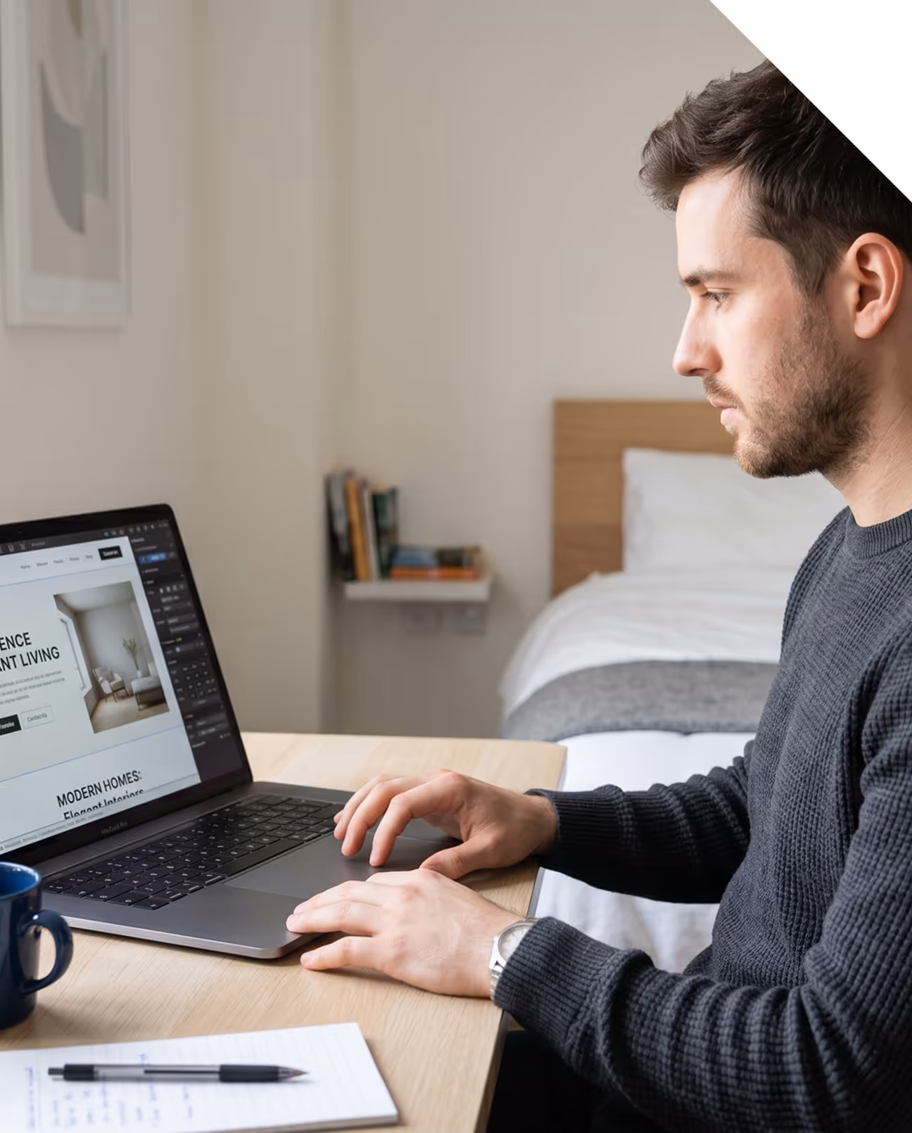 Homme concentré travaillant sur un ordinateur portable à une table avec un lit en arrière-plan.
