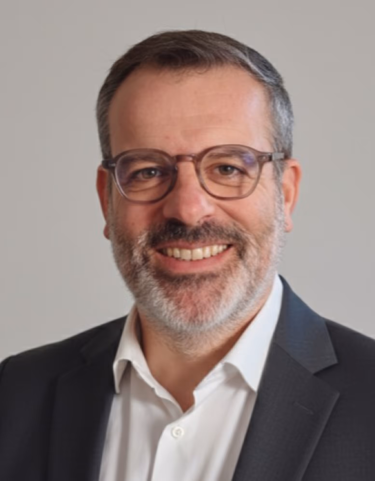 Homme souriant avec lunettes, barbe grise, chemise blanche et blazer noir sur fond neutre.
