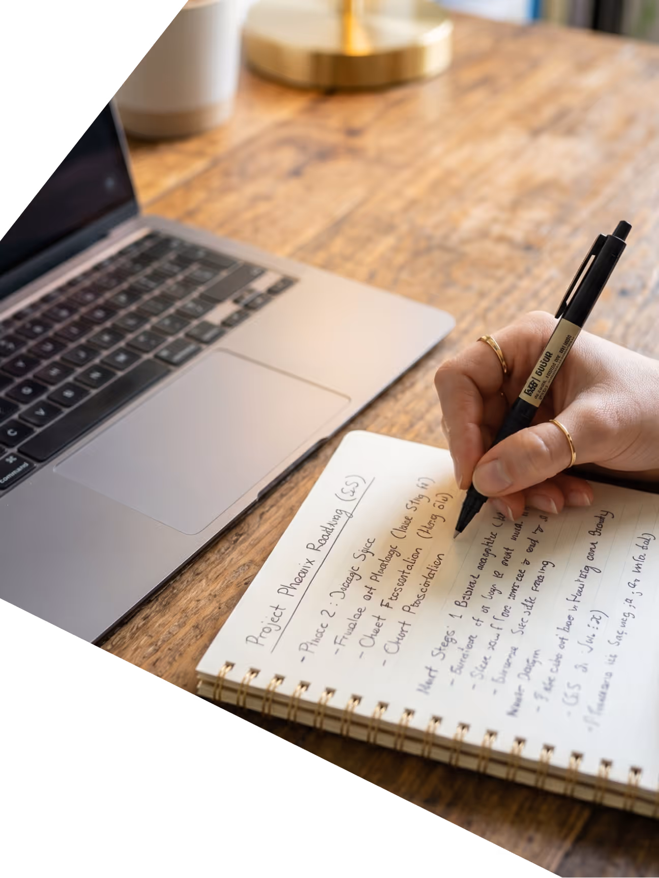 Person writing notes in a spiral notebook next to a laptop on a wooden desk.