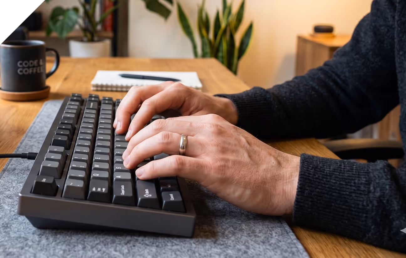 Une personne tapant sur un clavier mécanique noir sur un bureau en bois avec une tasse à café à côté.