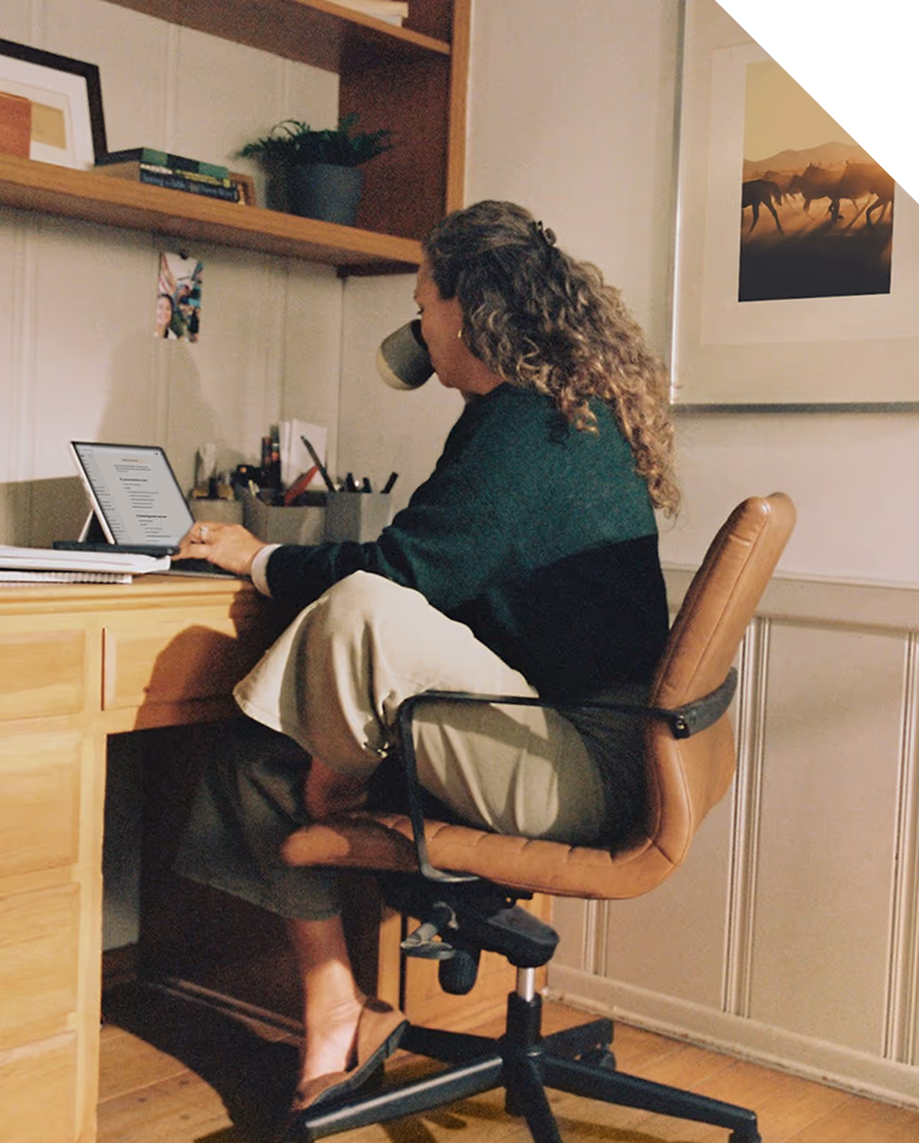 Femme assise à un bureau en bois, buvant dans une tasse et regardant un écran d'ordinateur portable.