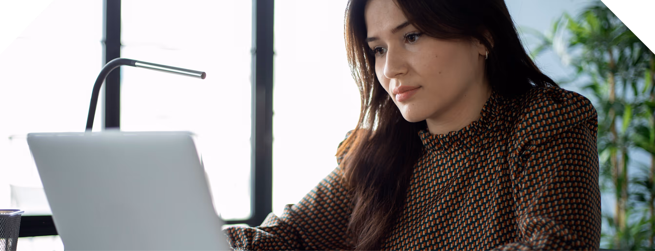 Une femme concentrée travaillant sur un ordinateur portable dans un bureau lumineux avec une plante verte.
