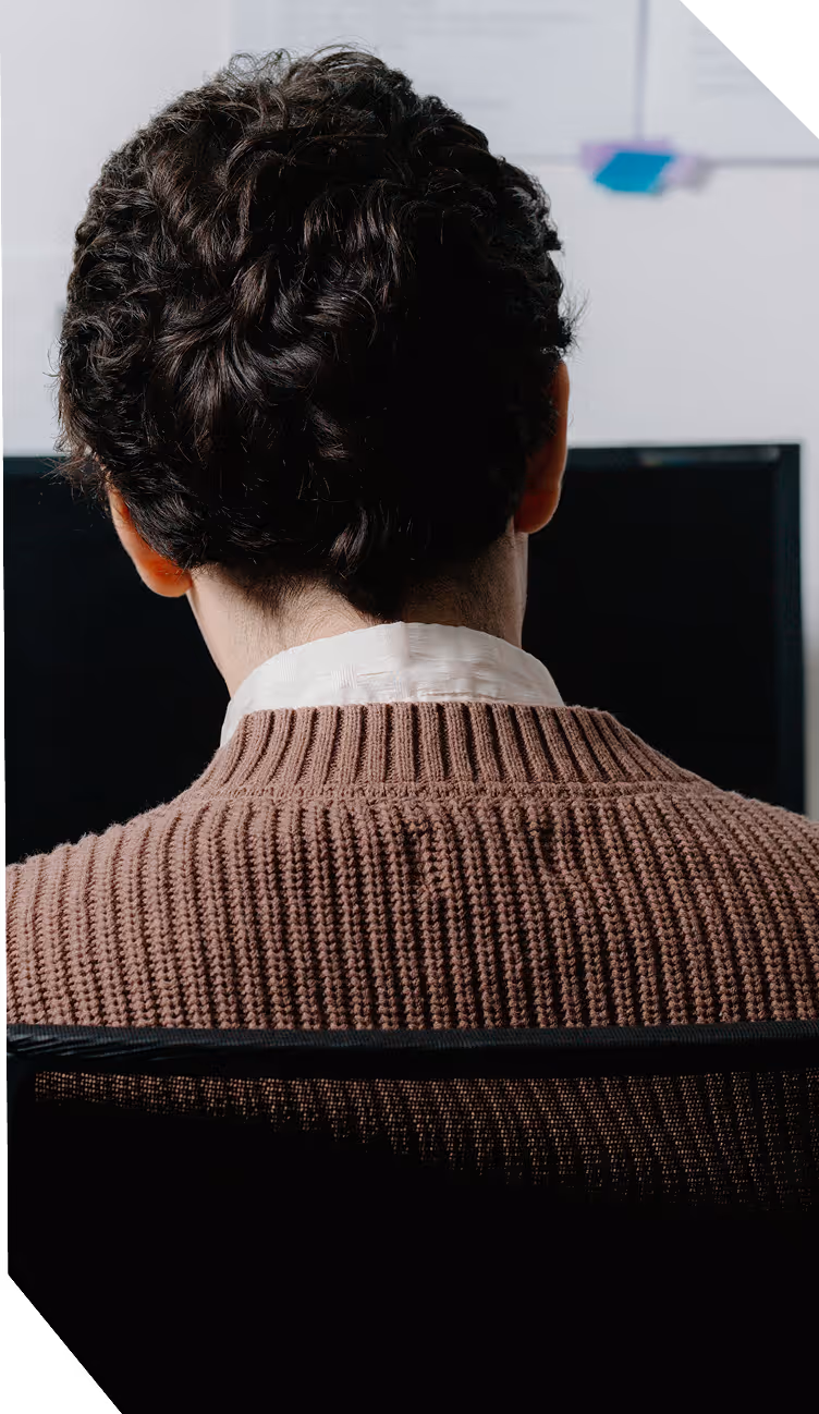 Homme vu de dos avec un pull marron tricoté assis devant un écran d'ordinateur.